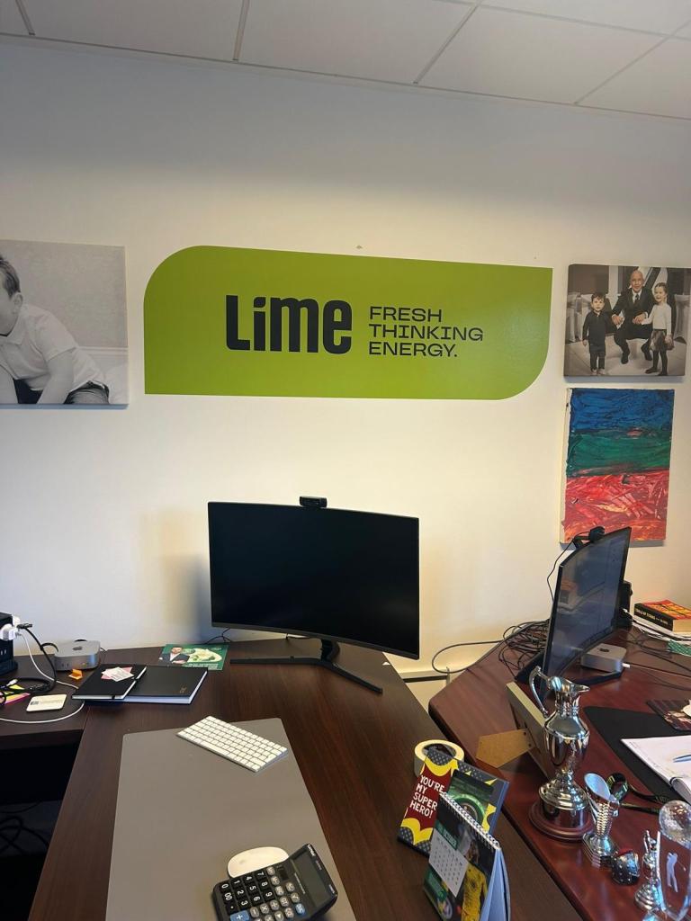 An office space featuring a green wall sign that says 'Lime FRESH THINKING ENERGY.' A desk with items including a computer monitor, a calculator, notebooks, and decorations can be seen in the foreground.